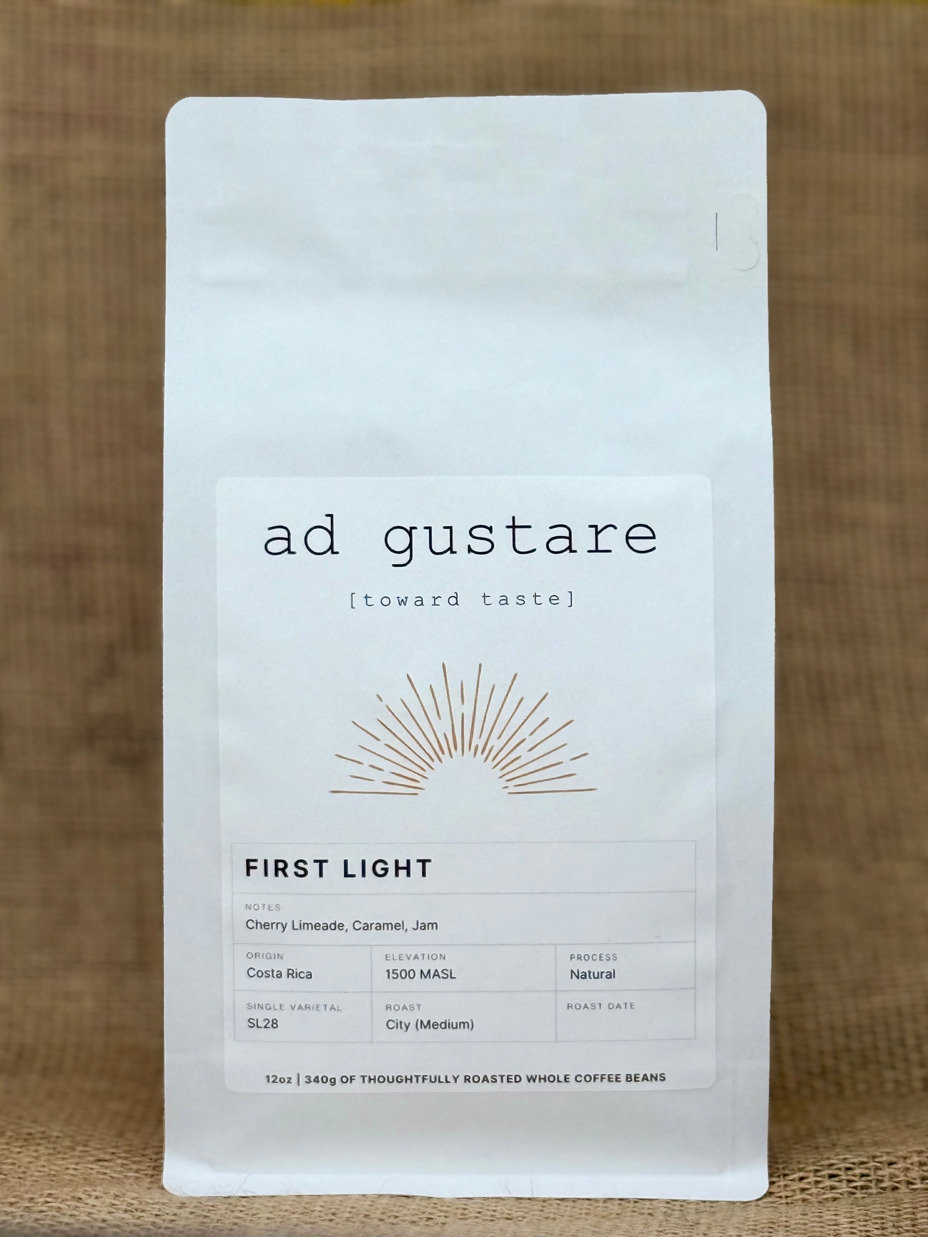 Ad Gustare First Light Costa Rica coffee bag with minimalist white label featuring a sunburst illustration, photographed on burlap.