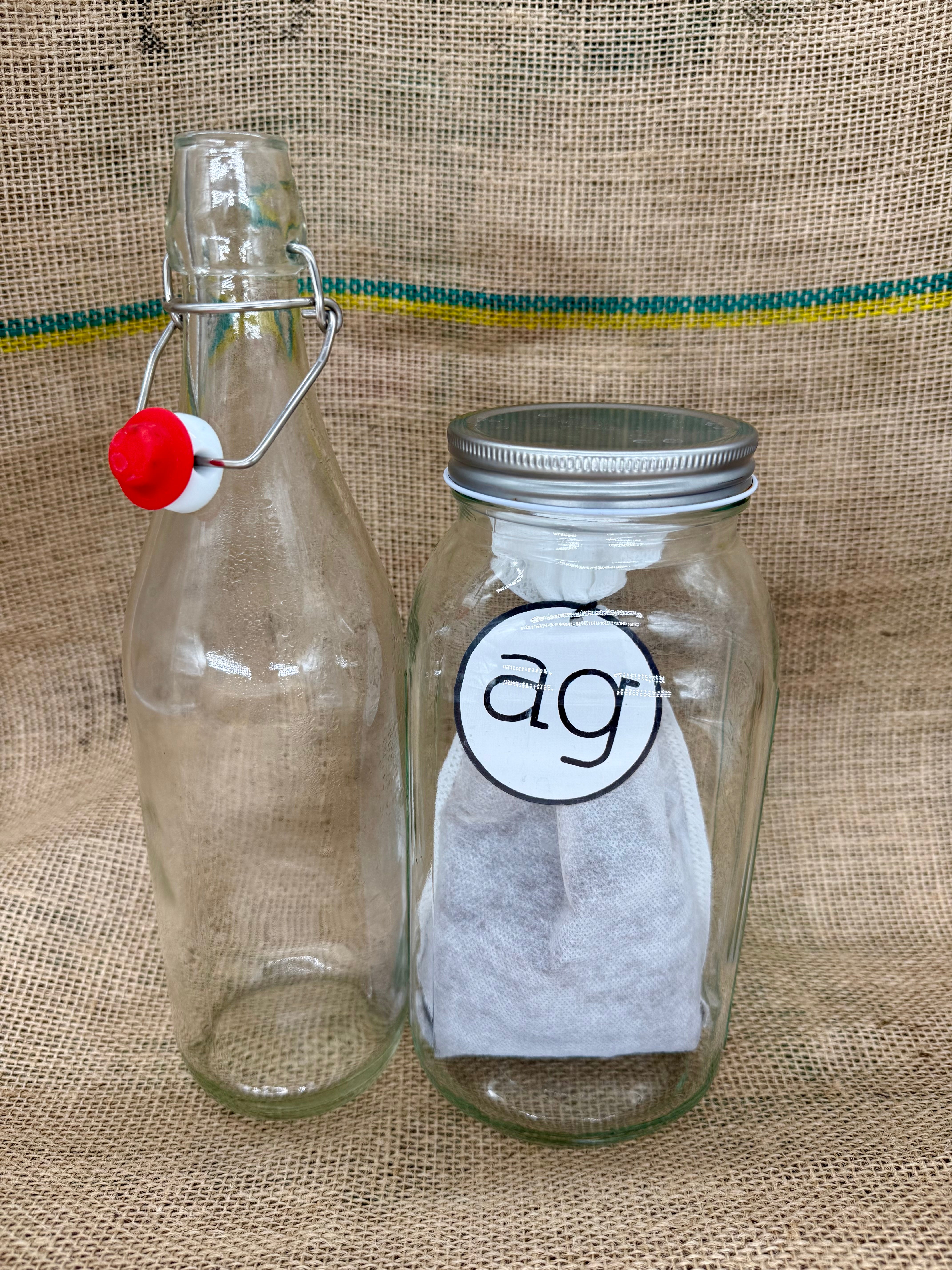 ad gustare single origin cold brew starter kit. Two glass jars with a red stopper and a metal lid on a textured fabric background. front view